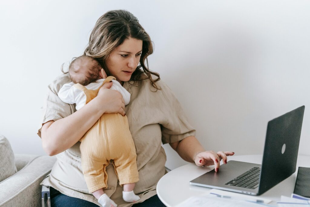 A woman holding a baby while looking for business ideas on a laptop